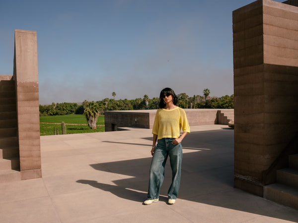 Une femme se tient debout au sein d'une maison mexicaine, elle porte une maille jaune et un jean large bleu