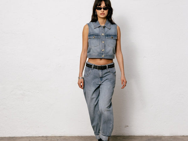 Une femme avance vers la caméra, habillée d'un ensemble denim à fines rayures blanches, elle porte des bottes noires et des lunettes de soleil