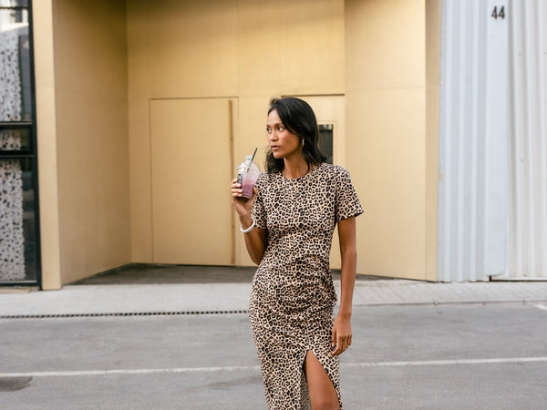Une femme se tient debout au milieu d'une ville, elle porte une robe midi léopard, moulante avec une fente sur le côté, des bottes mi-hautes et boit un café à la paille